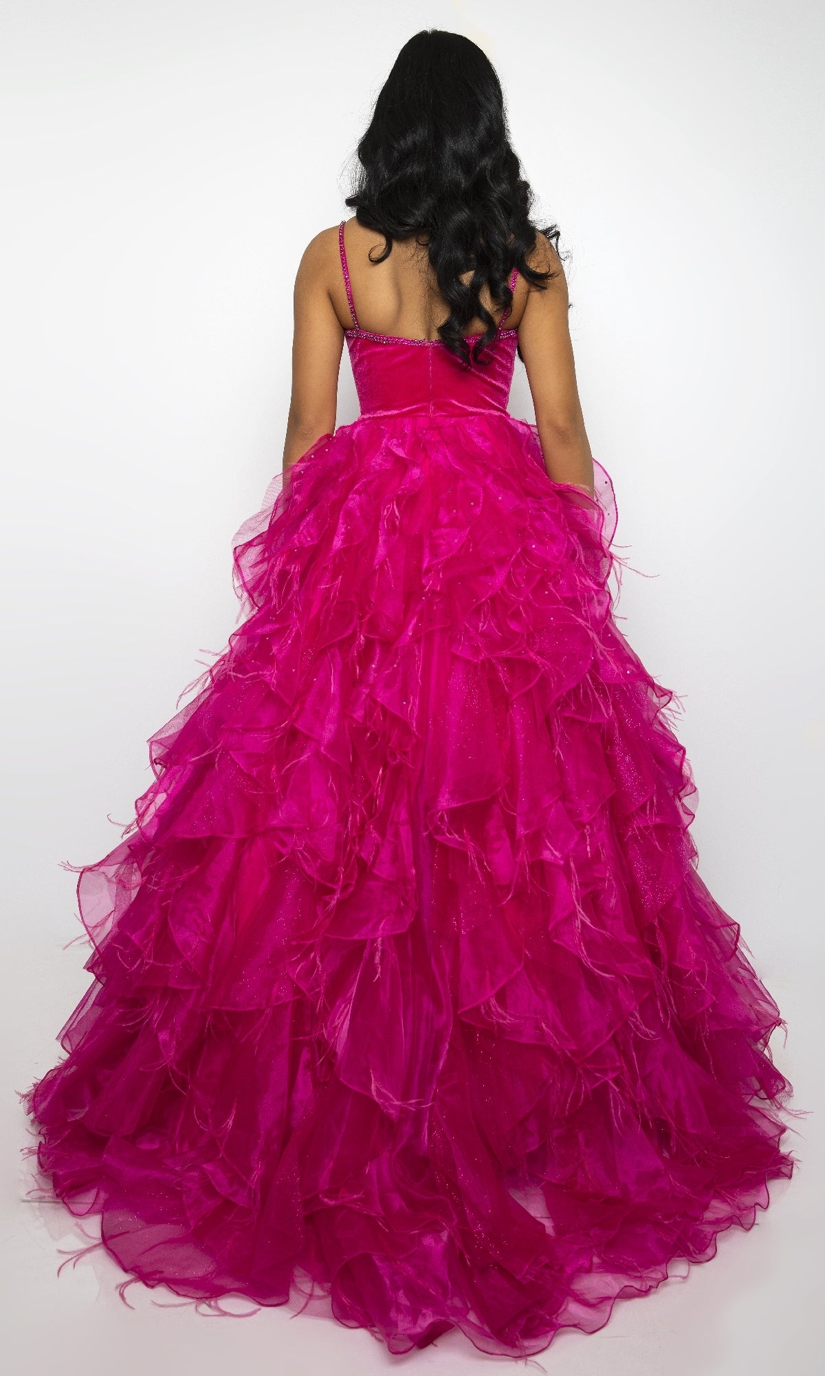 A person with long, dark, wavy hair is seen from behind wearing the Ava Presley 39554 long ruffled prom ball gown in bright pink, standing against a plain white background.